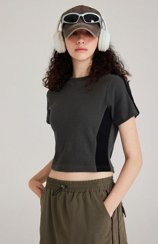 une fille au style normcore porte un t-shirt court basique noir et gris, un pantalon de survêtement vert foncé, un chapeau, des lunettes de soleil et un casque blanc