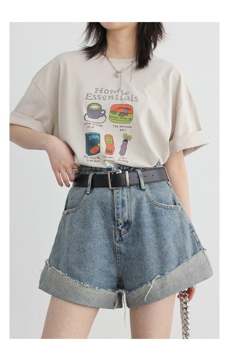 Una niña lleva pantalones cortos vaqueros desgastados y con aberturas, un cinturón negro, un collar plateado y una camiseta con estampado "Home Essentials".