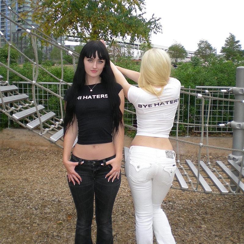 dos chicas vistiendo blusas de estética básica, la chica de pelo negro lleva una camiseta de hola enemigos en negro y la chica rubia lleva una camiseta de despedida de enemigos en blanco