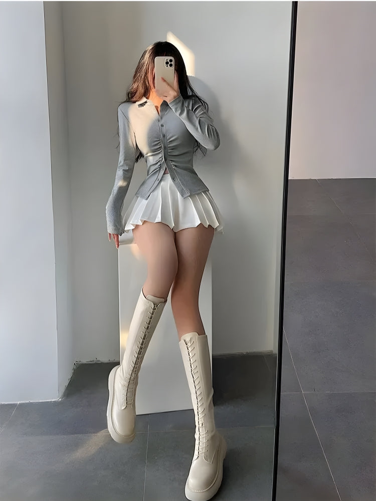 una chica con estética balletcore lleva una minifalda de tenis plisada blanca, un cárdigan gris con botones y botas militares color crema.