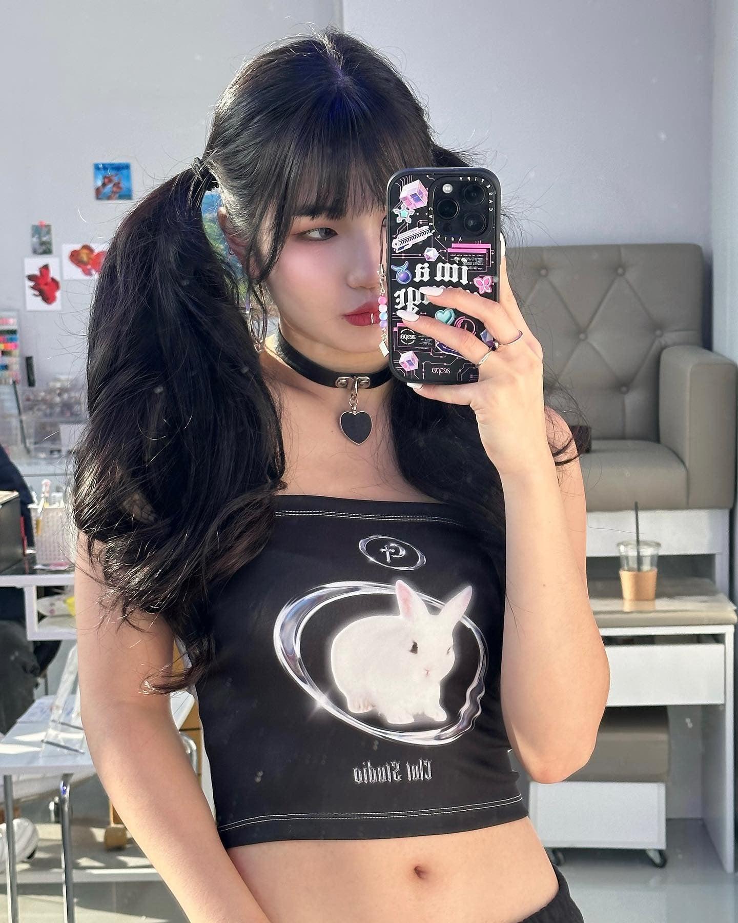 a girl is wearing a y2k rabbit tube top in black and a black choker with a heart shaped pendant on and she is holding her black phone taking a photo
